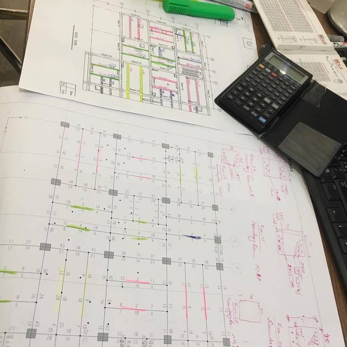 Printed structural drawings spread out on a desk, marked with pink and yellow highlighter, alongside a scientific calculator, keyboard, and engineering reference tables.
