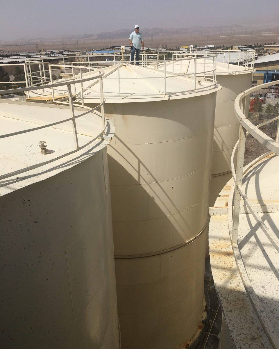 Construction worker on top of a large beige storage tank, showcasing the scale of the vertical vessels.