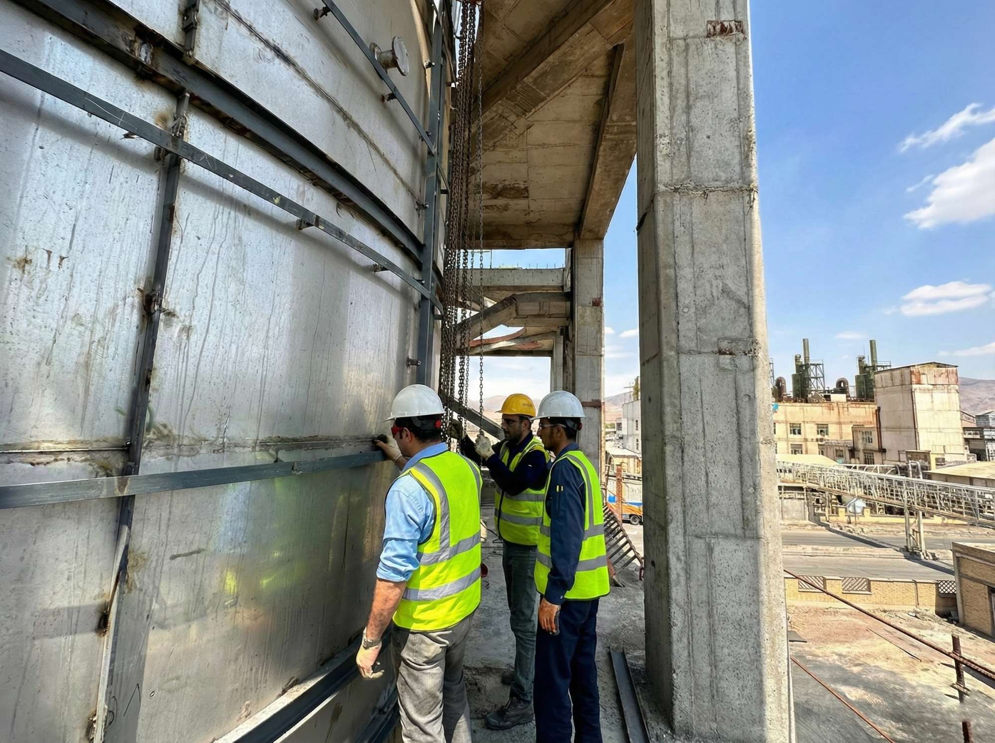 Three engineers in high-visibility vests and hard hats inspecting welds on a large vertical stainless-steel tank, standing on a concrete platform framed by reinforced concrete columns at an industrial plant.