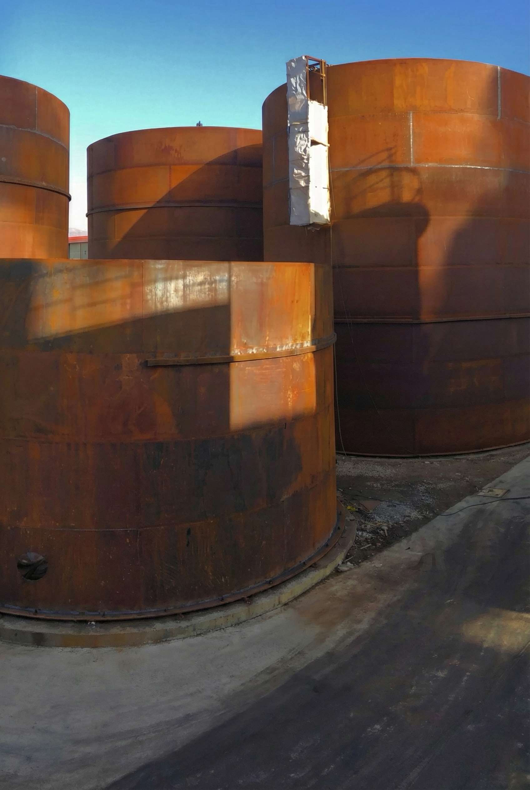 Several large cylindrical steel storage tanks with rust-colored surfaces stand outdoors on concrete foundations, casting long shadows under a clear blue sky