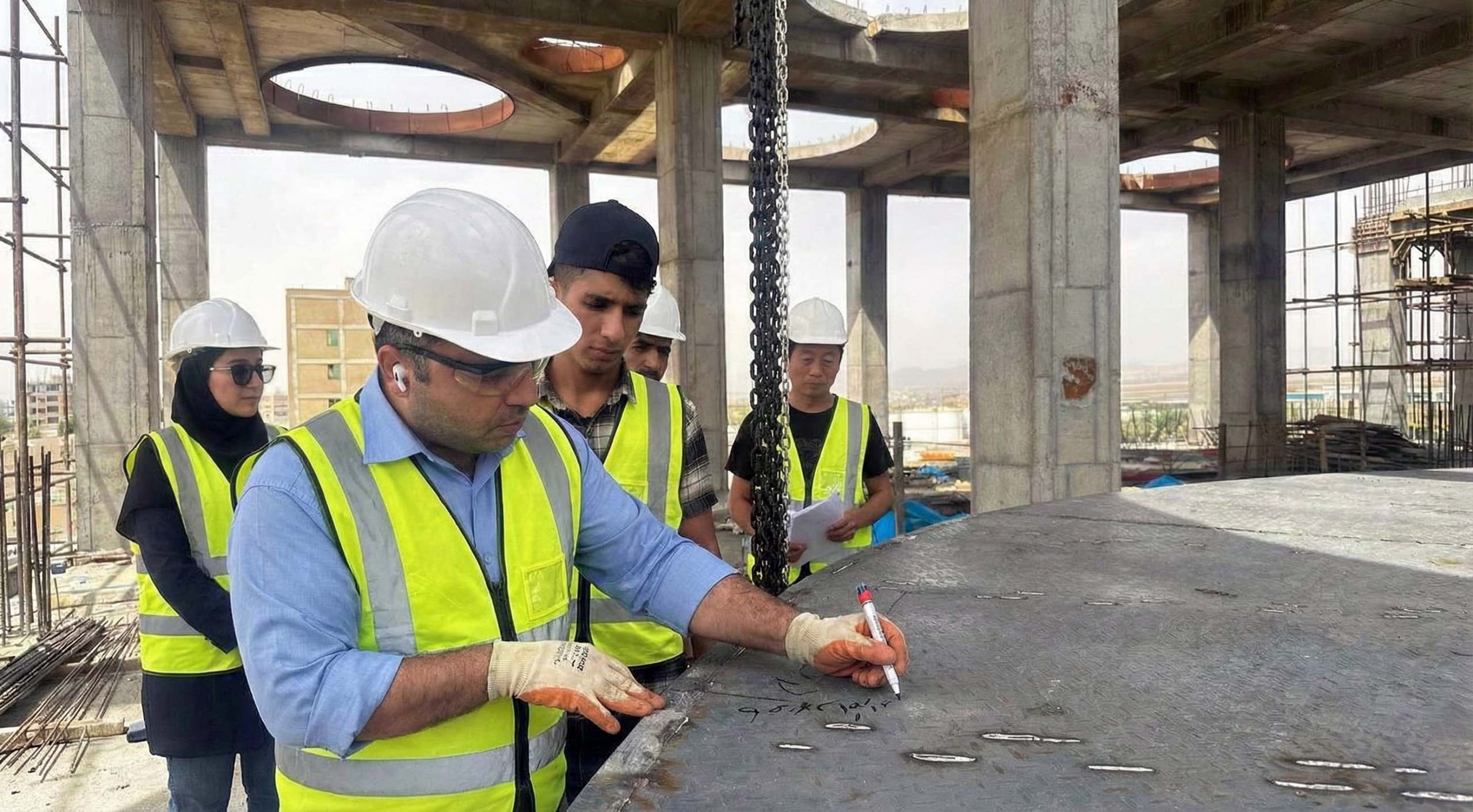 Close-up view looking across the top of a reinforced concrete slab under construction, with dense steel rebar grid and vertical bars protruding upward, while a worker in a hard hat stands in the background reviewing drawings on site.