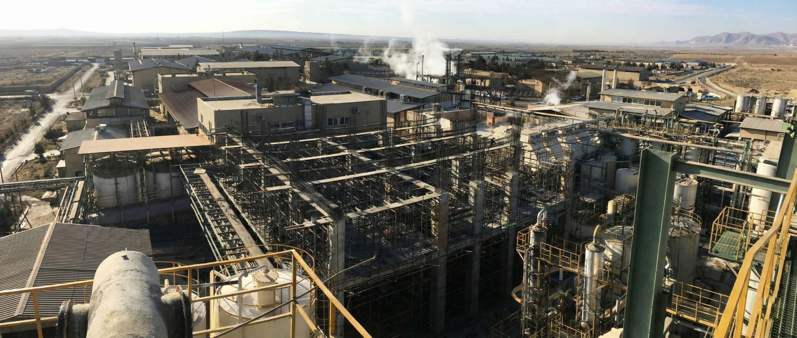Panoramic view of a large industrial plant with dense steel scaffolding, process equipment, and buildings, seen from an elevated platform with distant fields and mountains in the background.