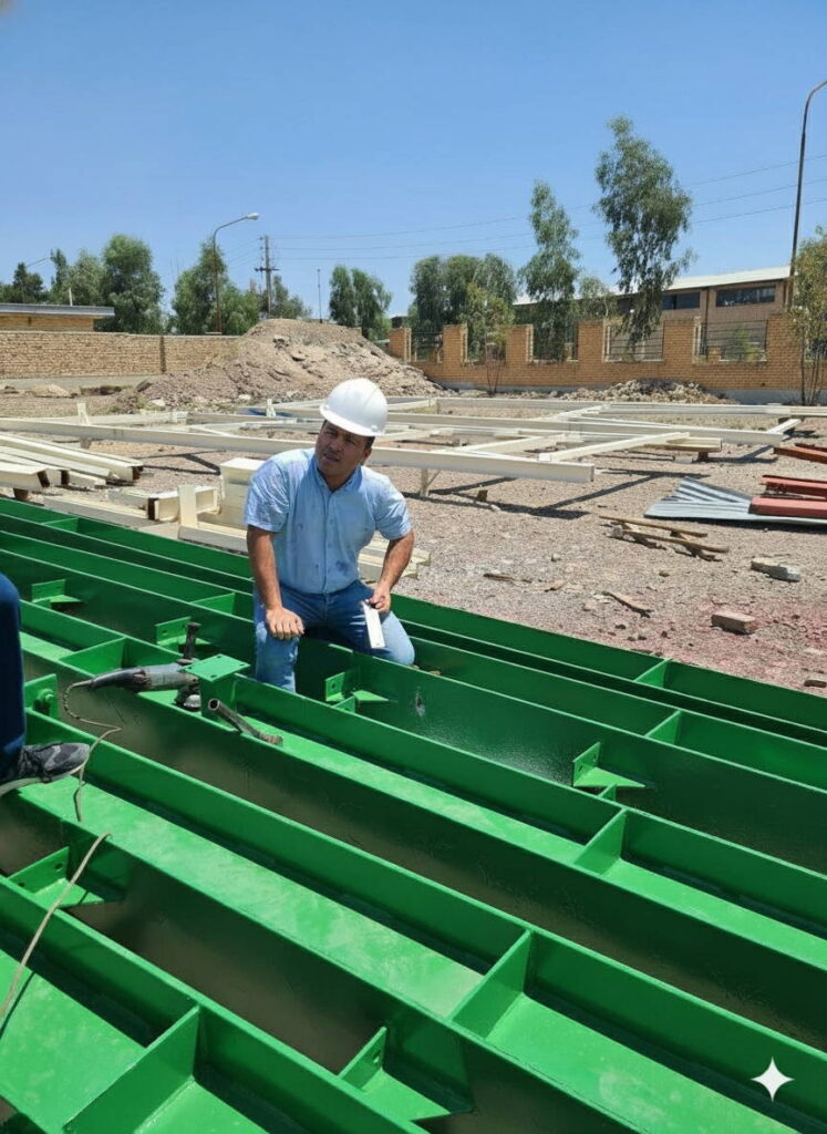 Engineer wearing a white hard hat and blue shirt inspecting green painted steel beams laid out in a fabrication yard.
