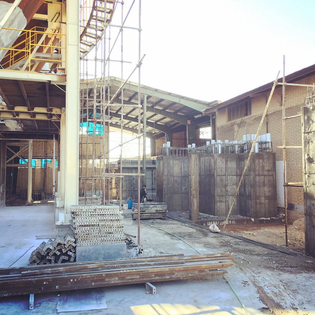 Wider view of industrial hall showing new heavy concrete walls and foundations being built alongside steel columns, scaffolding, and formwork stacks.