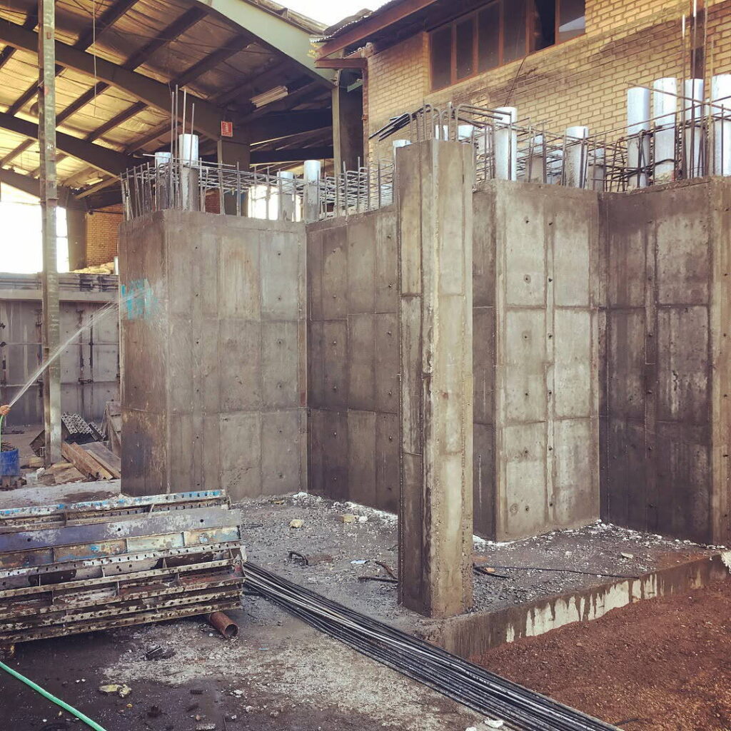 Massive reinforced concrete walls and bases under construction inside an existing industrial building, with exposed rebar extending from the top.
