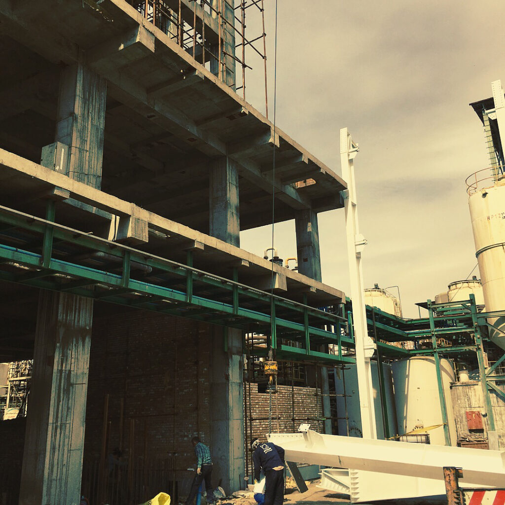 Workers assembling steel and concrete elements beneath a multi-story concrete frame with adjacent process pipe racks at the Industrial Complex Design project.