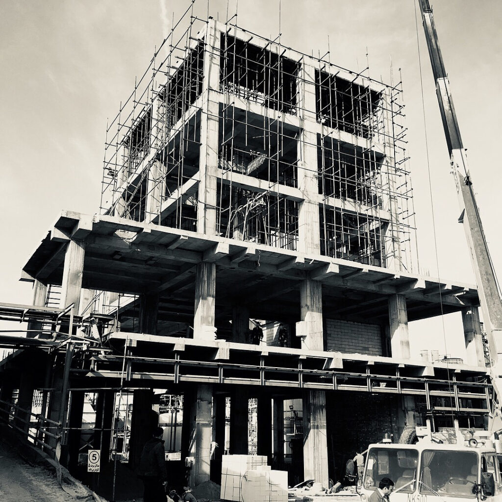 Black-and-white photo of the Industrial Complex Design process building under construction, with full scaffolding, crane, and workers around the concrete frame.