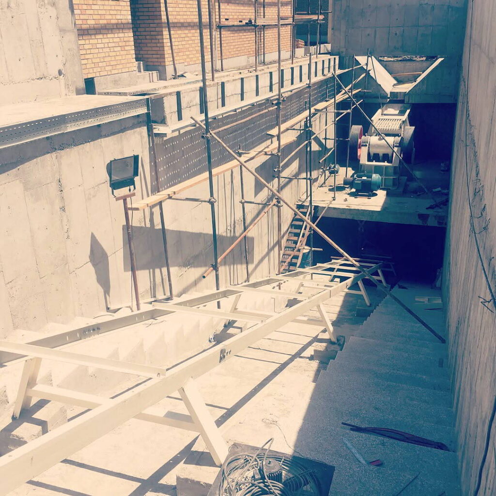 View down into a concrete trench with stairs and conveyor support steel, leading to a machine space at the far end.