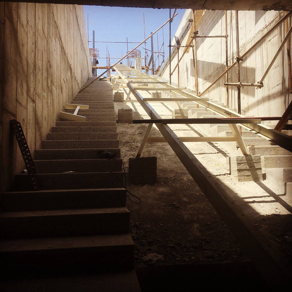 Steep concrete stair and adjacent sloped conveyor support corridor under construction between high retaining walls.