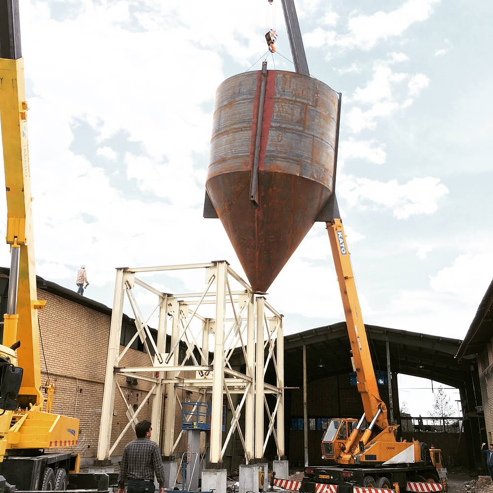 Cylindrical steel hopper being lifted by cranes into position above a cream-colored support frame between two industrial buildings
