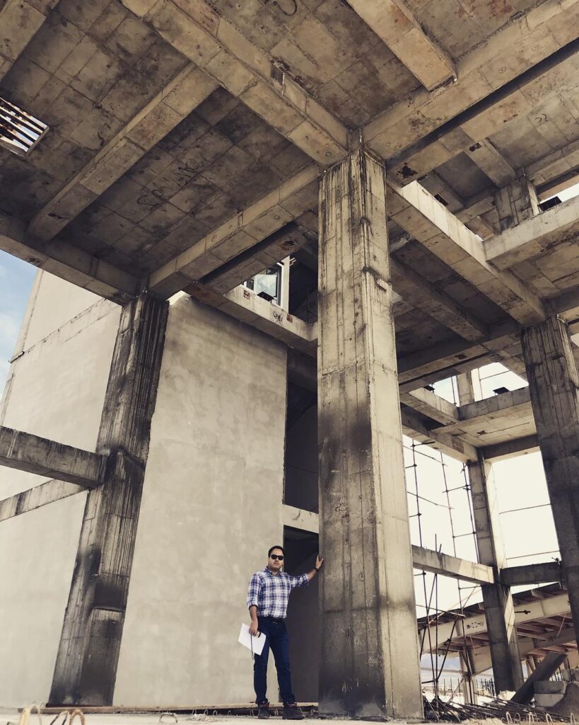 Engineer standing at the base of tall reinforced concrete columns inside an unfinished industrial building, assessing the exposed frame of the Industrial Complex Design project.