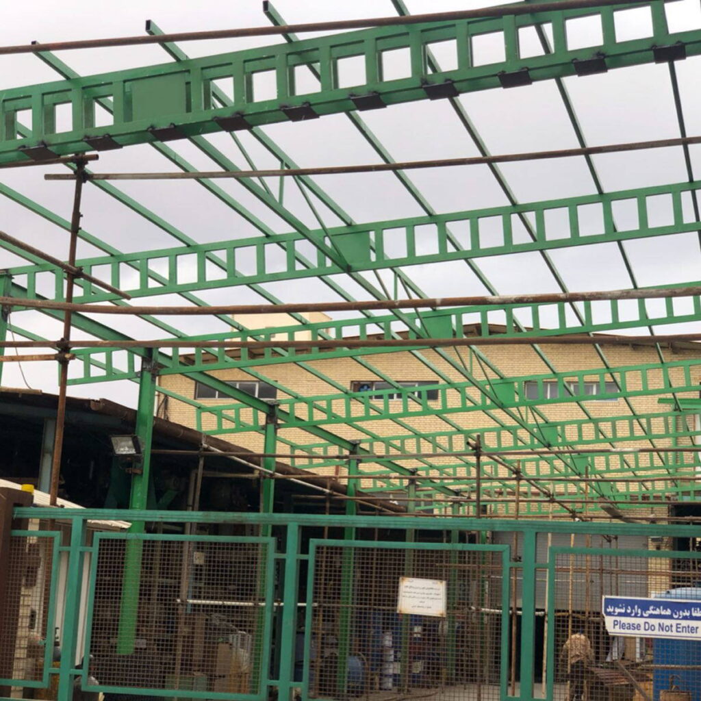Green lattice steel roof framing erected over an industrial area, supported by columns and surrounded by scaffolding.