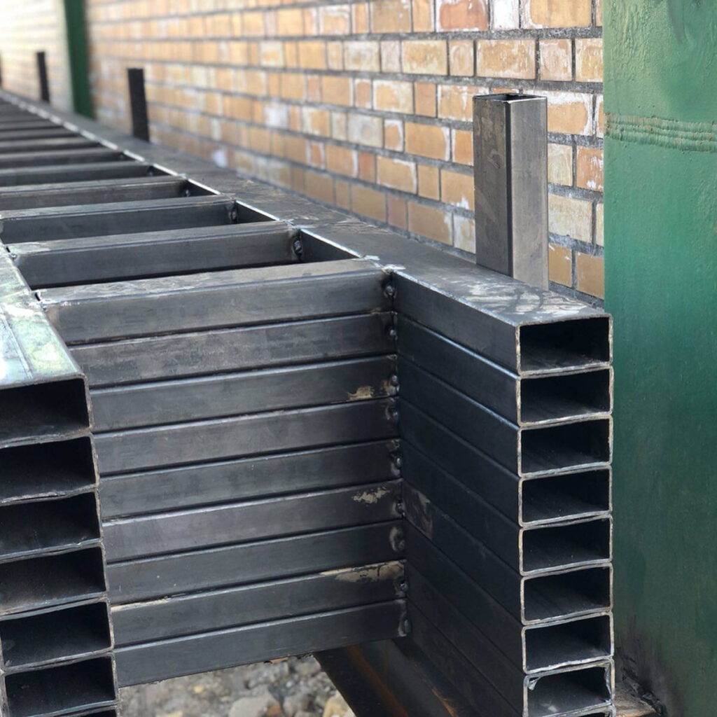 Close-up of stacked rectangular hollow steel sections welded into a box-like beam assembly alongside a brick wall.