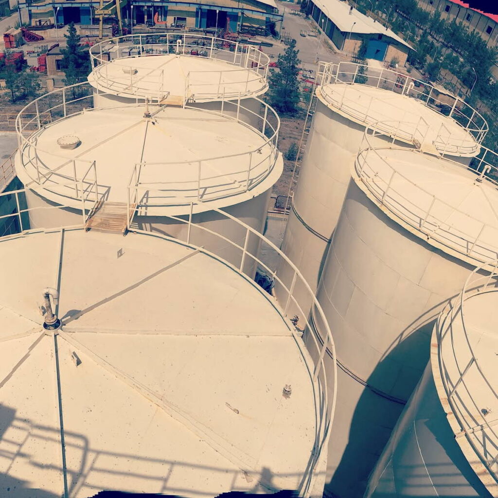 Aerial view of multiple large white cylindrical storage tanks with roof handrails and access walkways in an industrial yard.