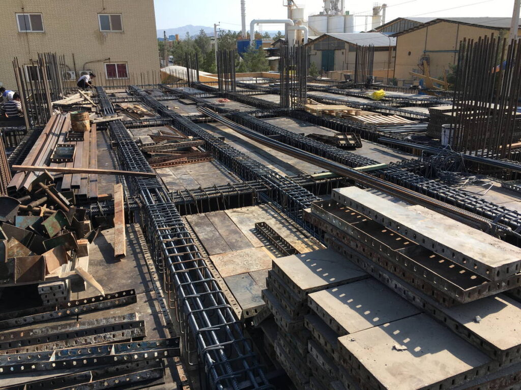 Wide view of an industrial building roof under construction, with continuous rebar beams, formwork panels, and stacked steel and concrete elements arranged across the deck.