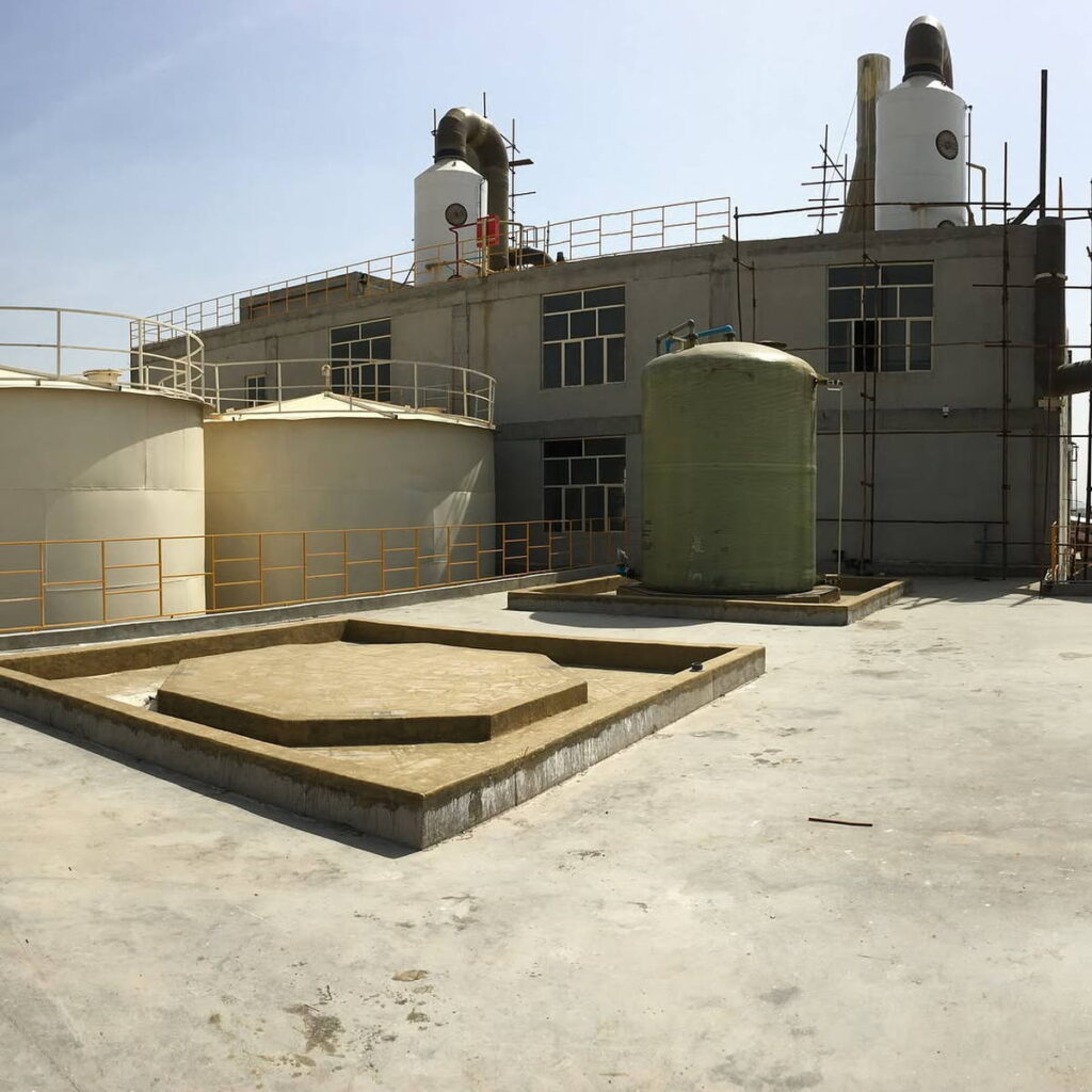 Chemical plant rooftop with circular storage tanks, a vertical fiberglass tank, and raised concrete foundations in front of a gray process building with scrubber towers.
