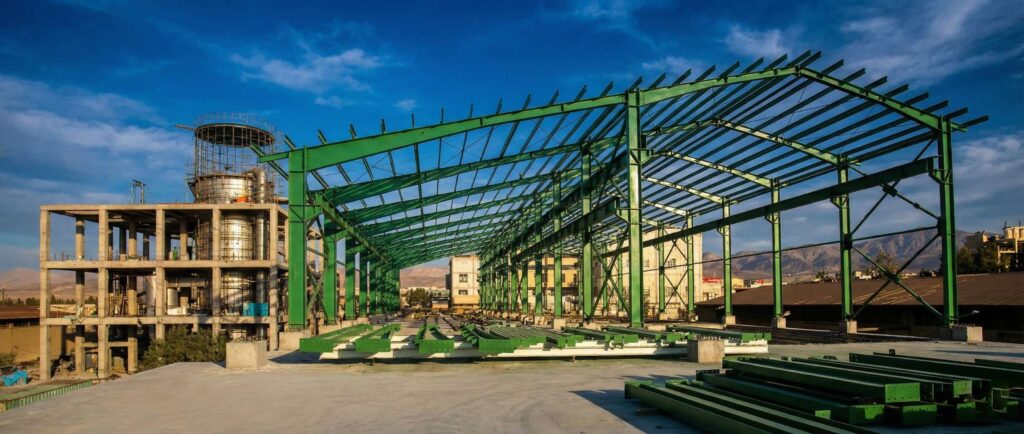 Wide-angle view of an industrial construction site with a large green steel warehouse frame rising beside a multi-story concrete process building that houses a vertical cylindrical tower, set against a blue sky and distant mountains.