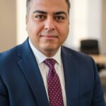 Professional portrait of Masood Keneshloo wearing a dark blue suit, light-colored dress shirt, and burgundy polka-dot tie, photographed in an office setting with soft natural light.​