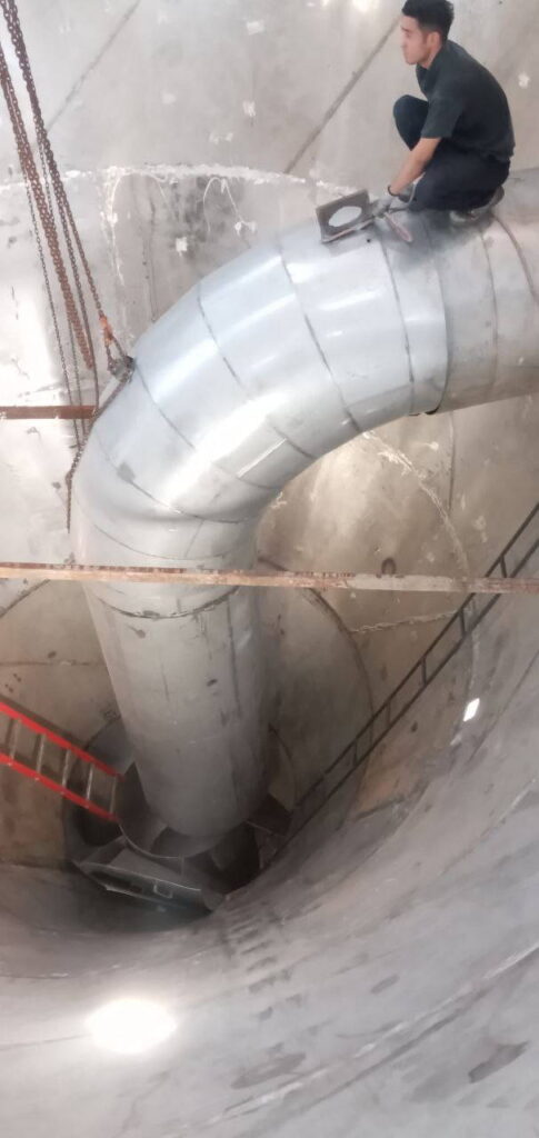 Worker installing a large metallic duct pipe inside a cylindrical reactor vessel during PAC production line construction.