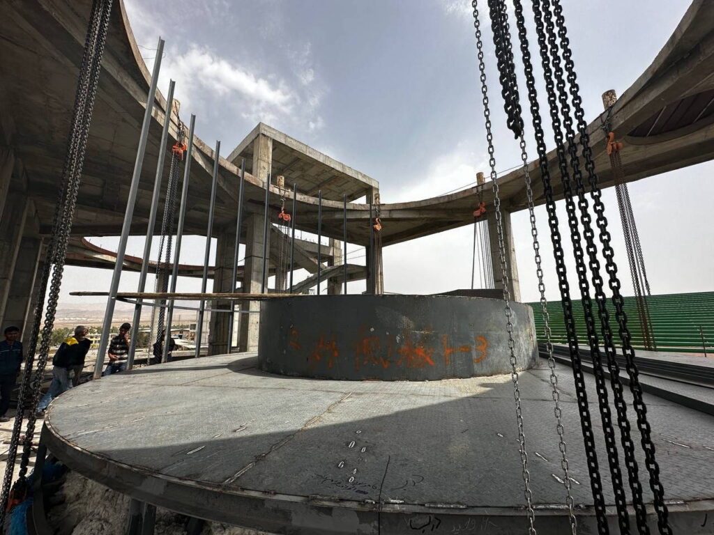 A large circular steel reactor base being hoisted by heavy chains at a construction site.