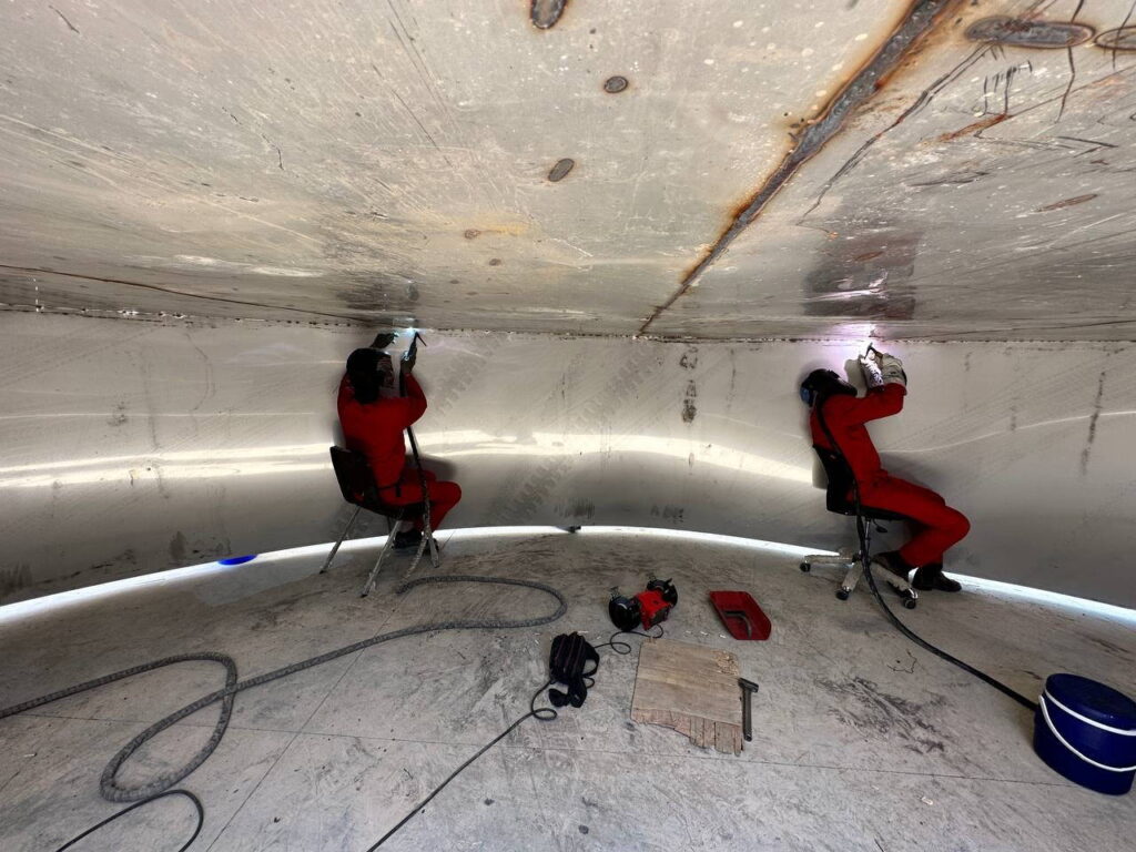Two welders in red suits performing seam welding inside a large stainless steel tank.