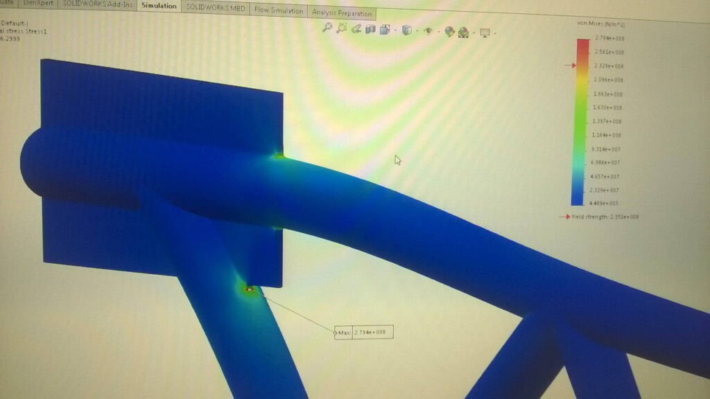 A SolidWorks simulation screen showing a heat map of stress distribution on a steel joint.