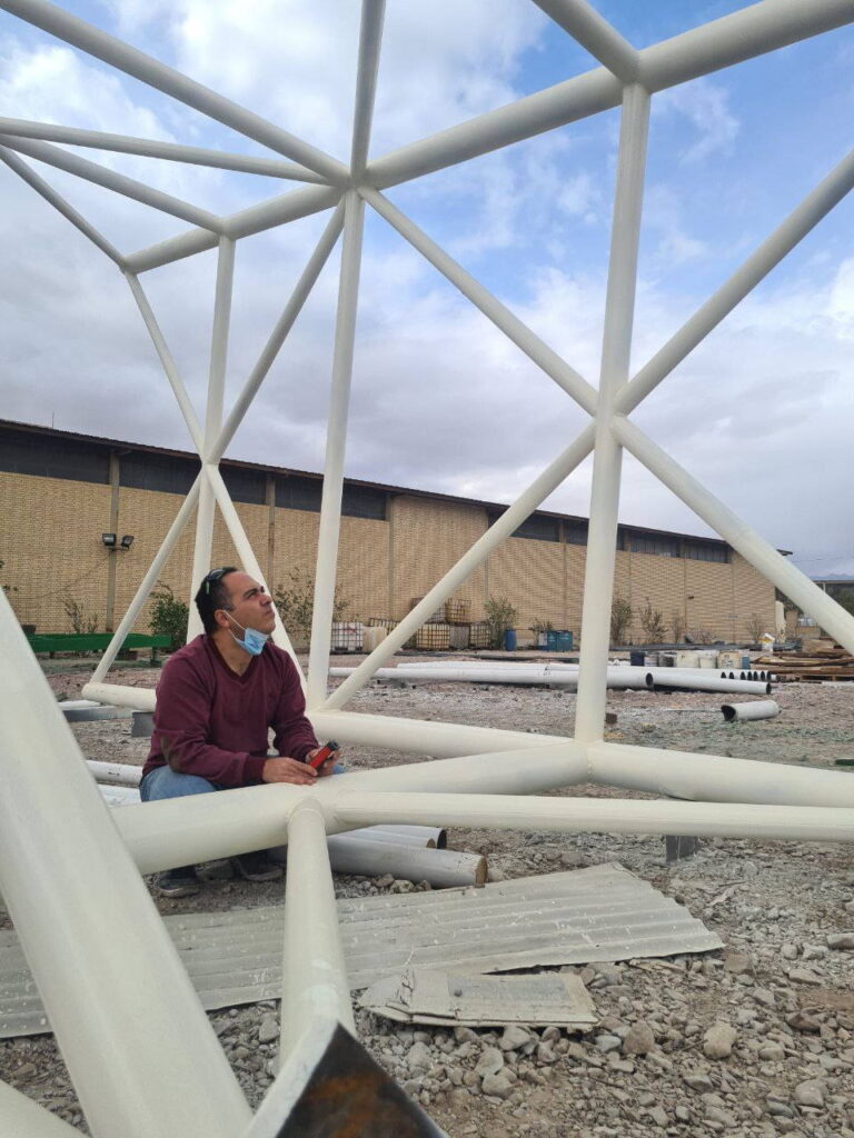 Masood Keneshloo kneeling inside a white triangular steel truss bridge on the ground during inspection.