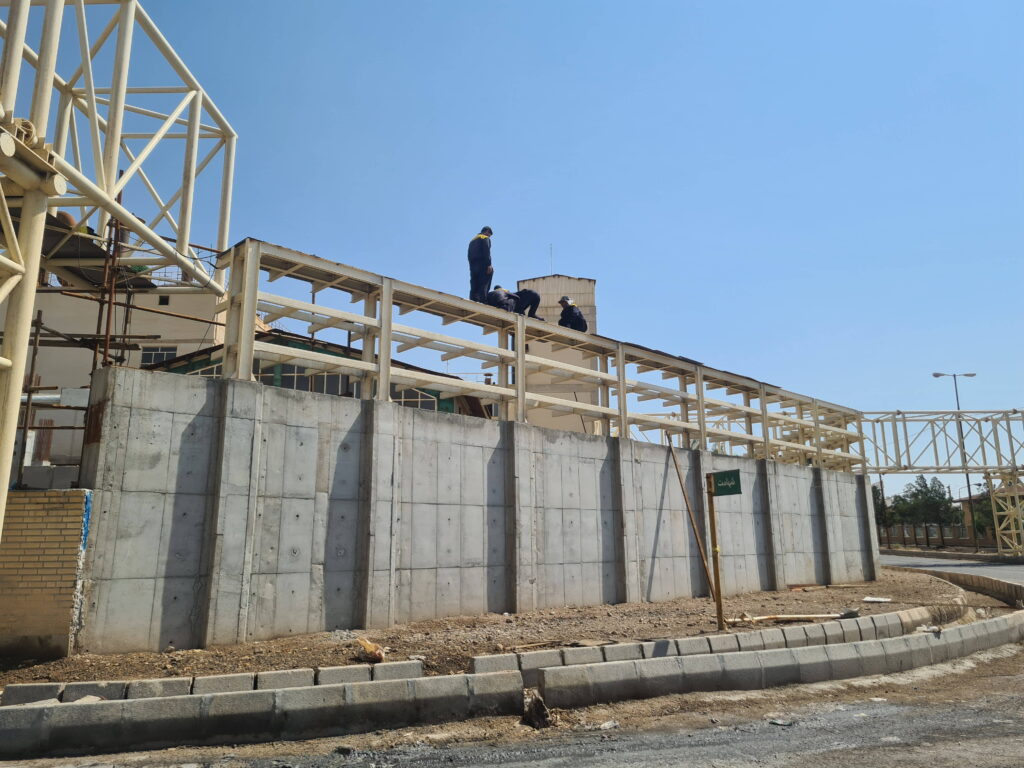 Workers installing a long horizontal steel pipe rack on top of a concrete retaining wall.