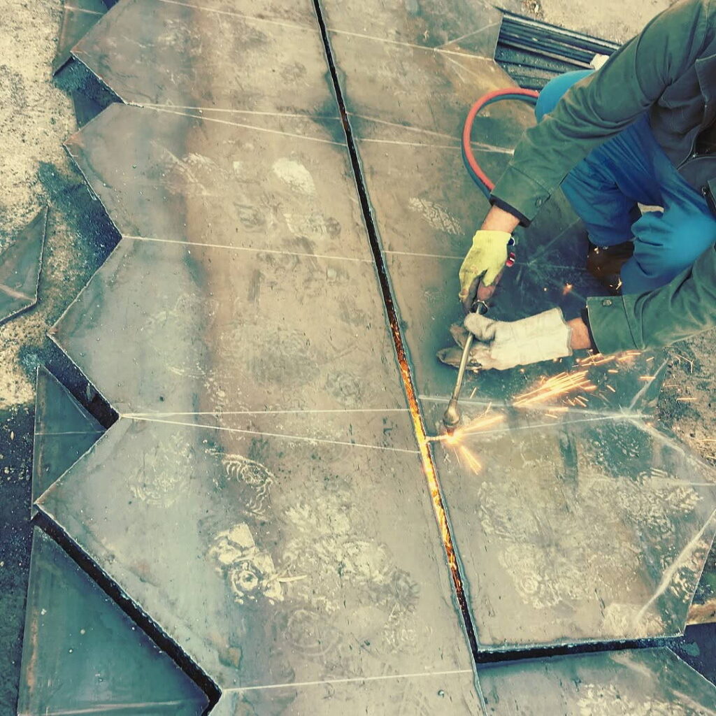 A worker using an oxy-fuel torch to cut complex geometric shapes out of a heavy steel plate.
