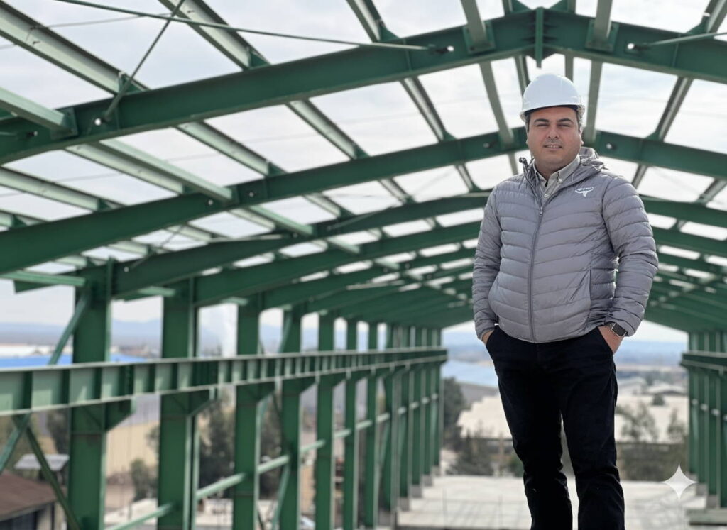 Masood Keneshloo in a white hard hat standing under the green rafters of a steel structure.