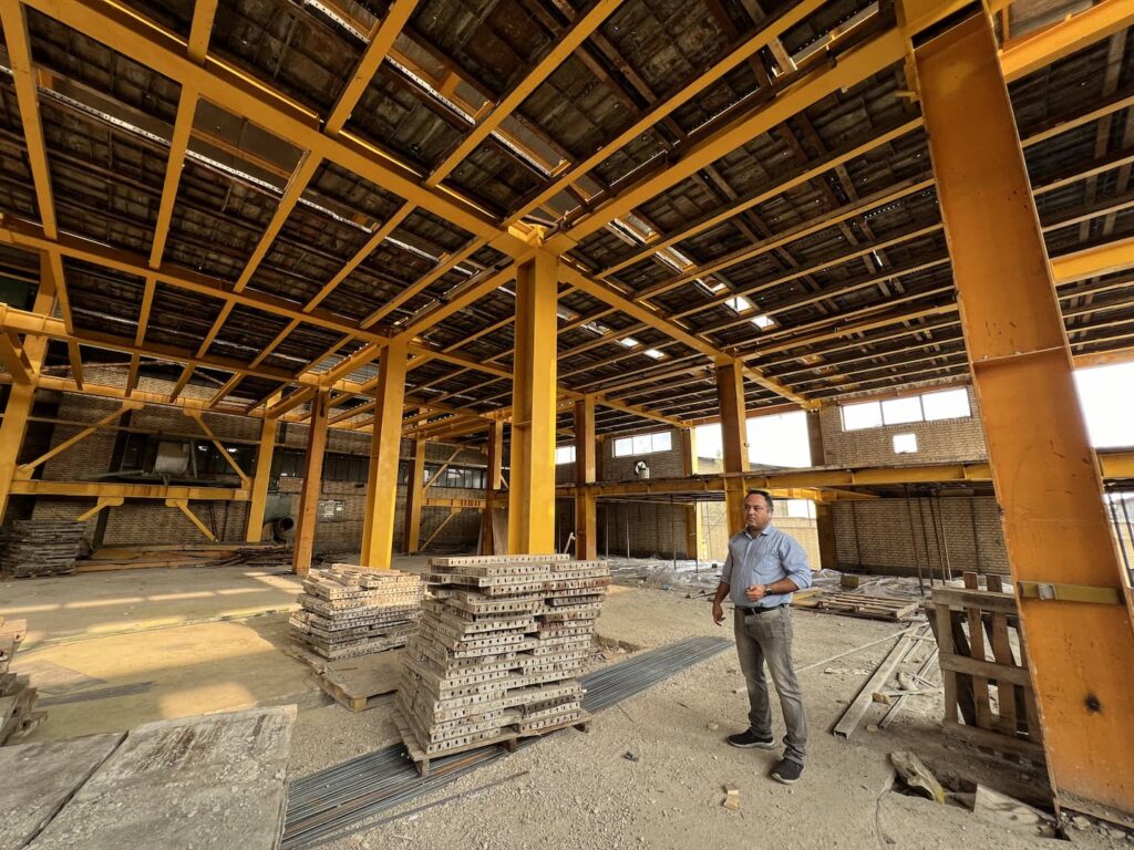 Masood Keneshloo standing inside a building with a dense yellow-painted steel ceiling framework.