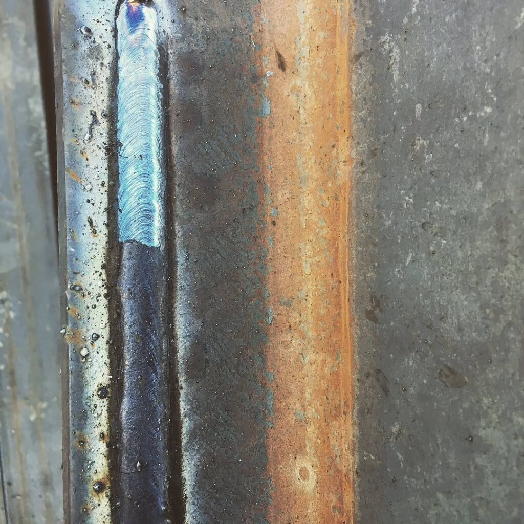 Close-up of a bright, silver-colored weld bead on dark steel with heat tint.