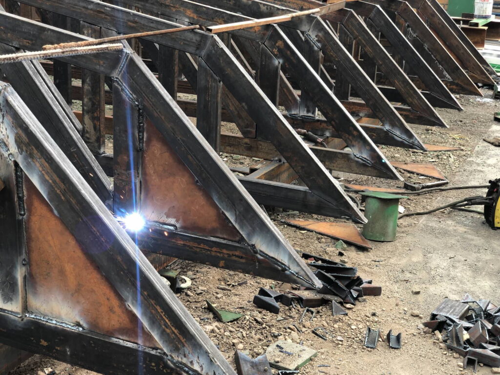 Arc welding of heavy triangular steel brackets arranged in a row on the ground, with a bright welding arc visible on one connection.