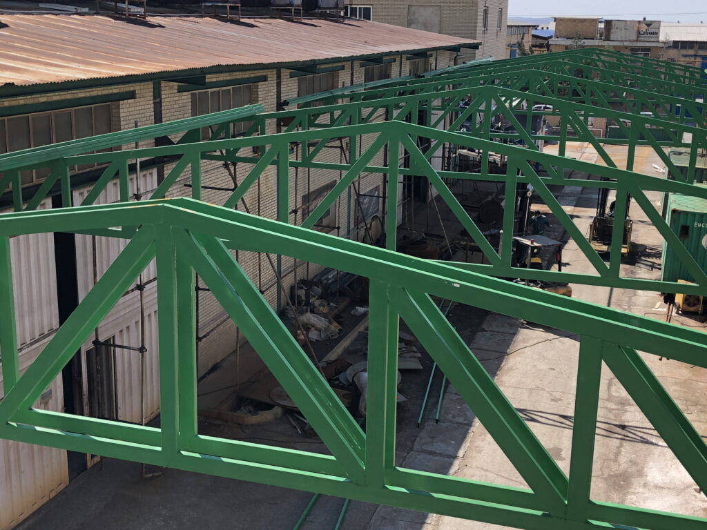 Row of green steel roof trusses installed along the side of an industrial workshop building.